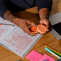 A person holds an open round orange Golden Hour pill tin containing orange Golden Hour, photographed from above on a wooden desk with an open notebook, pen, laptop, and sticky notes visible.