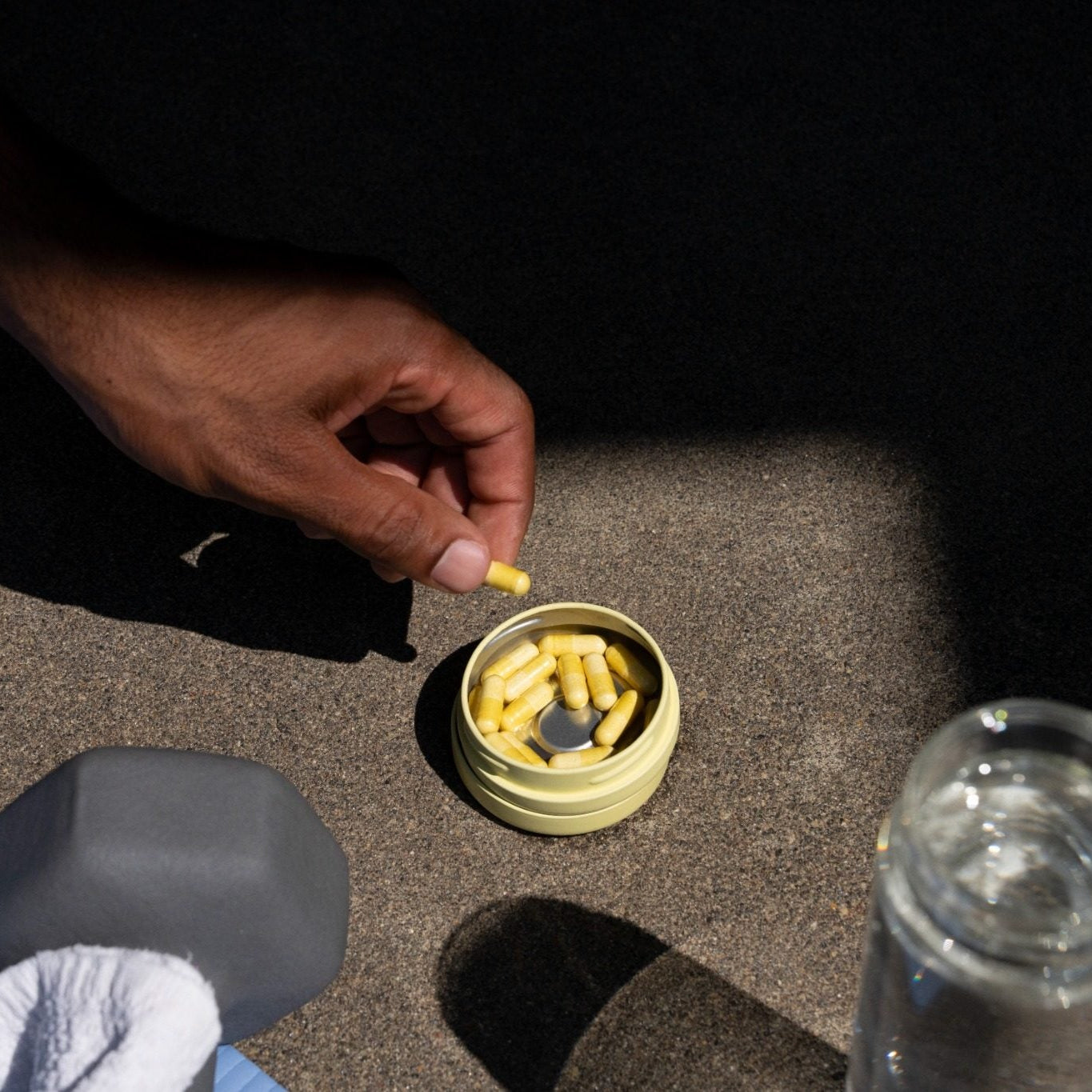 A hand picks a yellow Sunbeam capsule from the Sunbeam tin filled with more Sunbeam capsules, set on a dark textured surface with dramatic shadows. A clear bottle and a dumbbells are partially visible in the foreground.