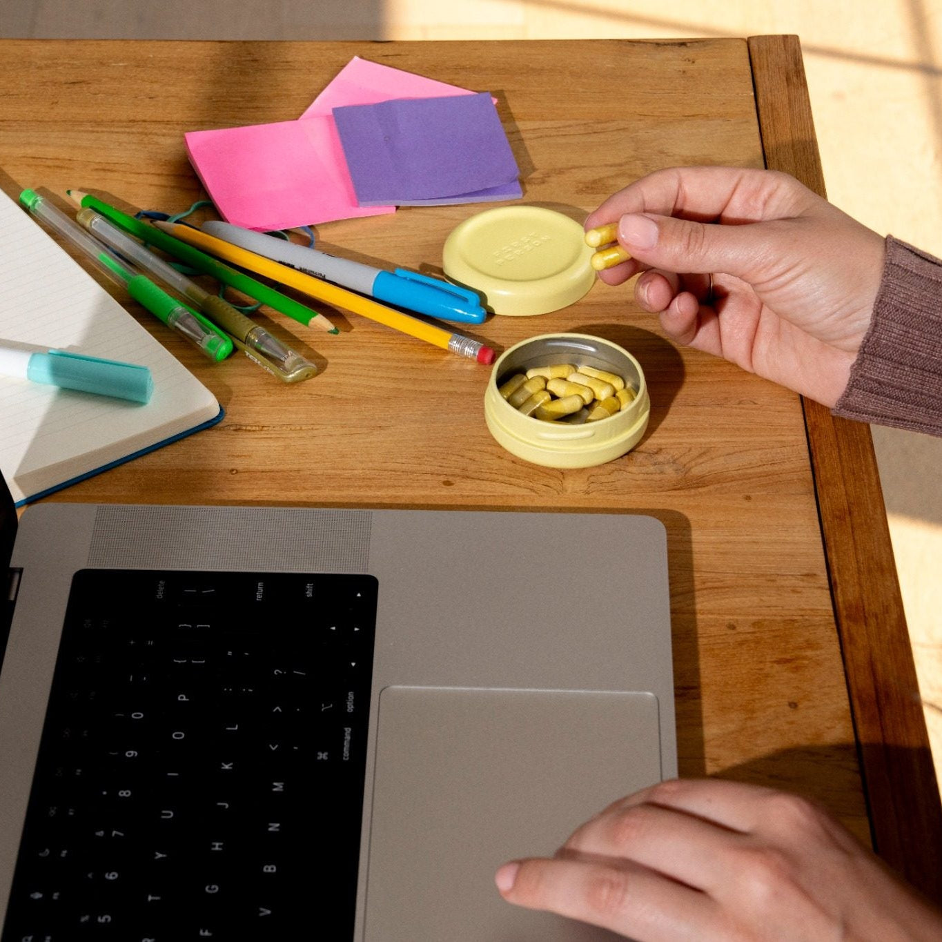 A person's hands holding two Sunbeam capsuleson a wooden desk next to an open laptop, with pens, sticky notes, and other stationery items in the background.