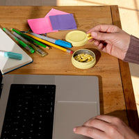 A person's hands holding two Sunbeam capsuleson a wooden desk next to an open laptop, with pens, sticky notes, and other stationery items in the background.