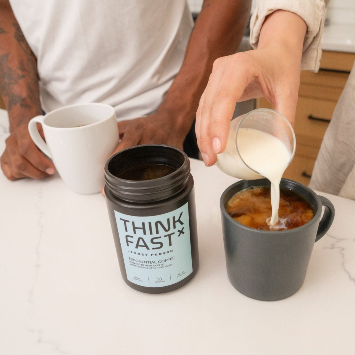A person pours milk from a small glass pitcher into a dark gray mug containing ThinkFast coffee, with a THINK FAST container.