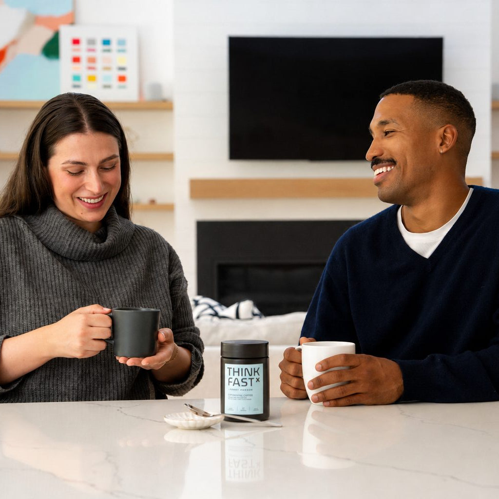 A smiling woman in a gray turtleneck holds a mug of ThinkFast coffee while sitting next to a man in a navy sweater holding a white mug with ThinkFast coffee. Between them on a white marble counter is a ThinkFast container.