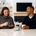 A smiling woman in a gray turtleneck holds a mug of ThinkFast coffee while sitting next to a man in a navy sweater holding a white mug with ThinkFast coffee. Between them on a white marble counter is a ThinkFast container.